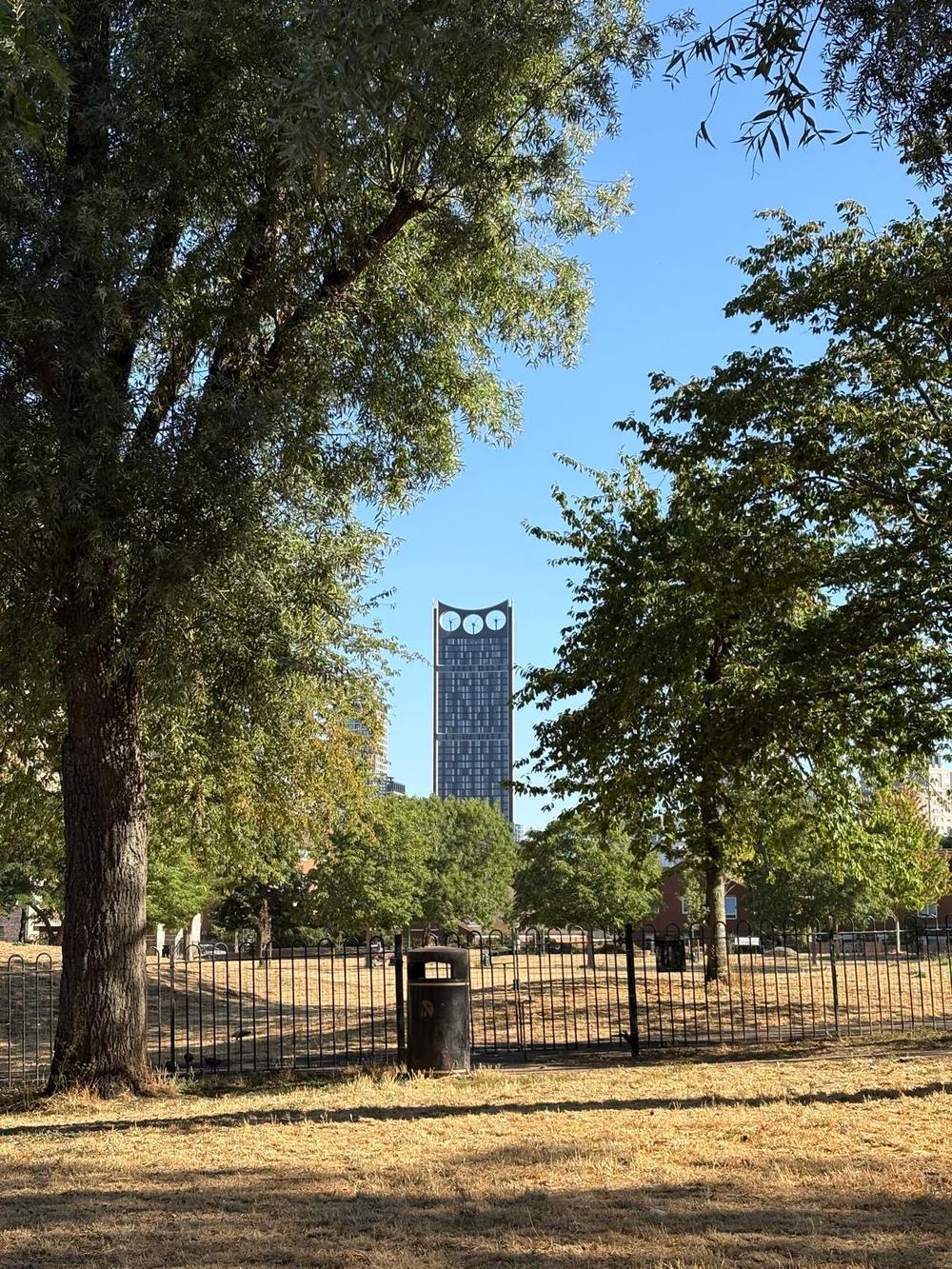 The Strata tower seen between two trees from a park with scorched grass, iron railings and a municipal bin in the foreground.